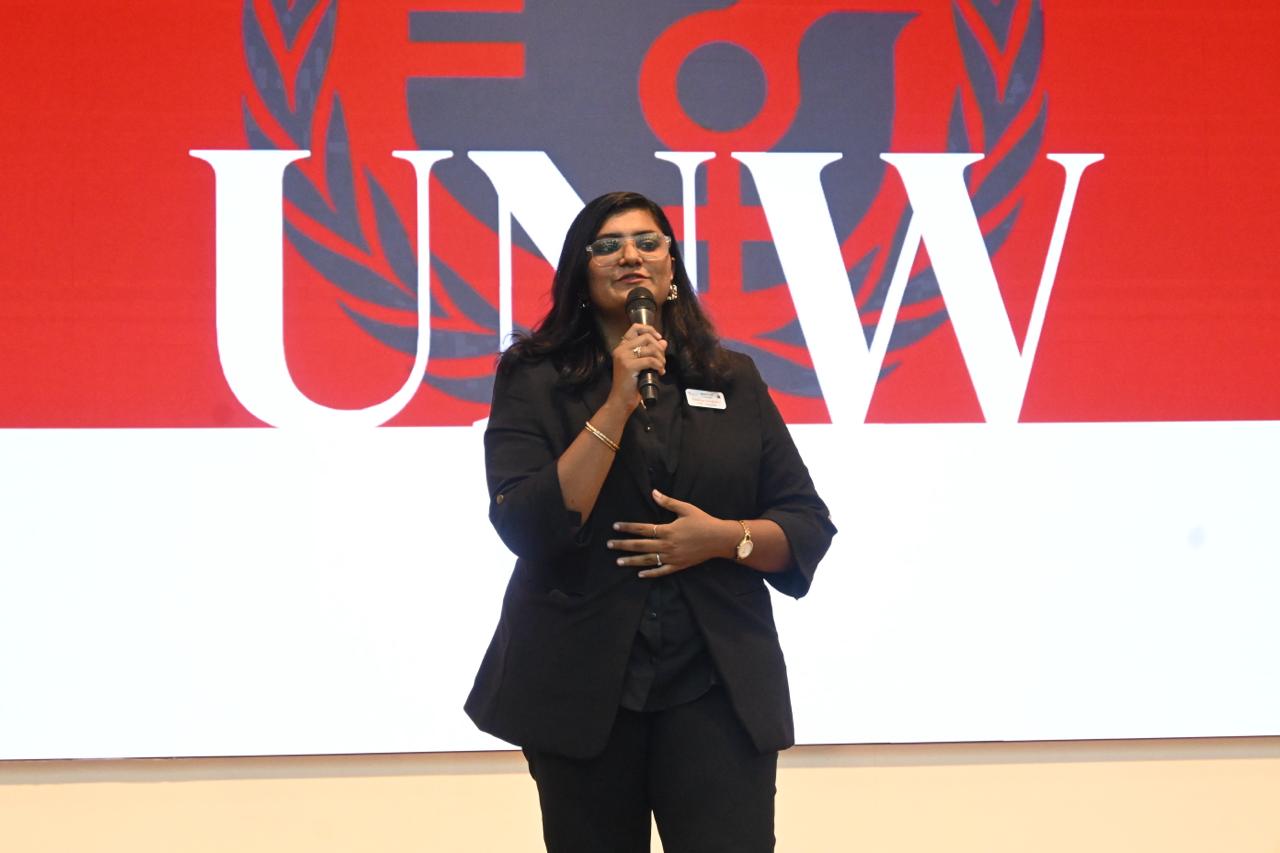 Frishthaa Dawulath speaking into a microphone on stage in front of a screen showing the UN Women (UNW) logo.